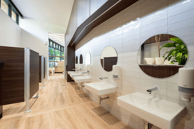 A bright contemporary communal bathroom with a long row of wall-mounted white rectangular basins, chrome fixtures, round mirrors, and white rectangular wall tiles. Dark timber privacy partitions line the left side, while light timber-look flooring, high clerestory glazing, and a dramatic dark timber ceiling beam add warmth and architectural interest. A leafy plant is visible in one mirror reflection.
