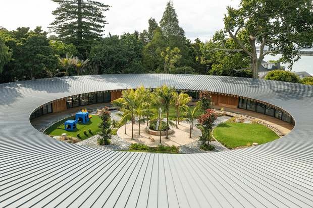 Aerial view of a low, circular or oval building organized around a landscaped central courtyard. The broad ribbed metal roof forms a continuous ring with a large elliptical opening, while timber-lined glazed rooms face the courtyard. The courtyard contains palms, gravel paths, lawn, planting beds, and a small blue outdoor play or seating area. Dense mature vegetation surrounds the property, with a glimpse of water and neighbouring buildings beyond.