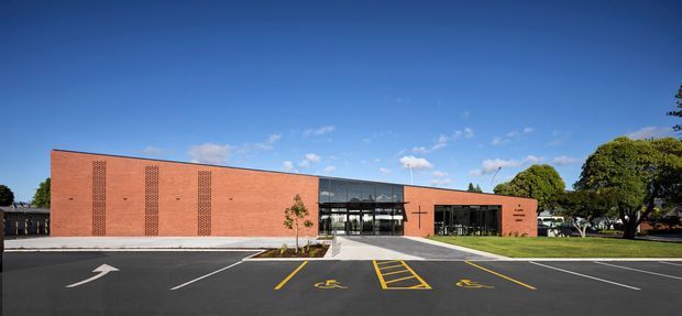 A low, single-storey contemporary institutional or commercial building with a long red-brick facade, regularly spaced vertical brick detailing, and a central glazed entrance. The building fronts a freshly paved parking area with marked accessible parking bays, landscaped lawns, mature trees, and a clear blue sky.