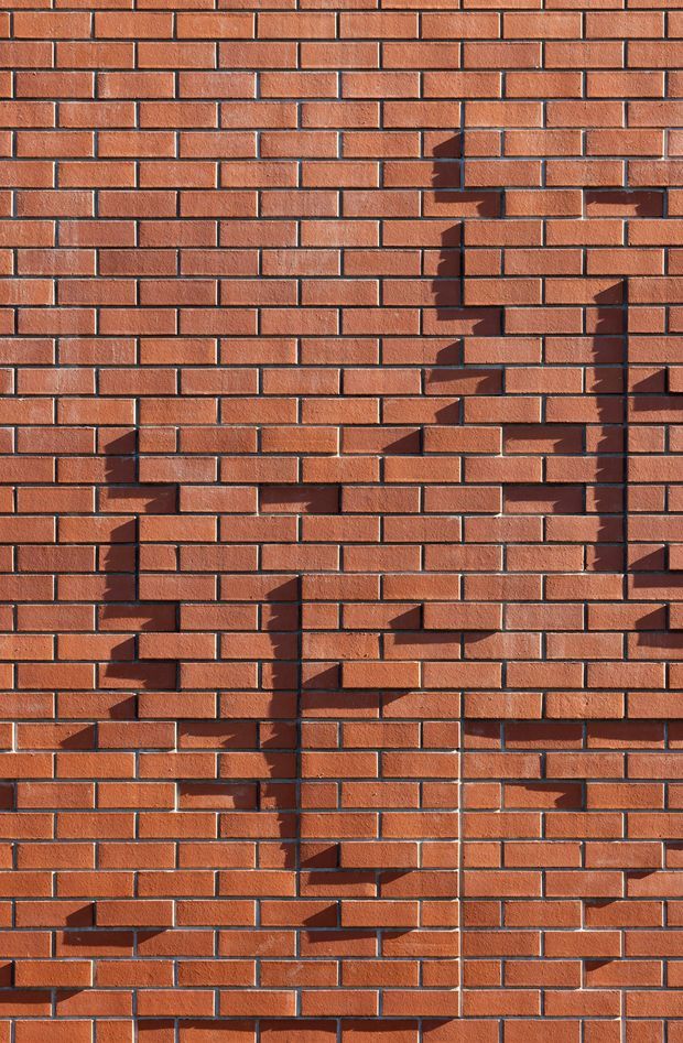 Close-up view of a red-orange brick wall in a regular running-bond pattern, with light mortar joints and strong sunlight casting angular shadows from projecting brick details across the facade.