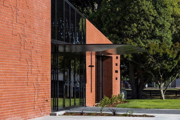 Exterior view of a contemporary brick building entrance, featuring red brick walls, tall dark glazed panels, and a projecting flat metal canopy. The entrance opens onto a paved walkway bordered by lawn and mature trees.