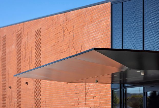 Close-up exterior architectural detail showing a contemporary entrance canopy projecting from a red brick facade. The canopy has a dark metal soffit and warm copper-toned metal upper surface, with tall dark framed glazed panels and a glazed entrance visible at right. Decorative brickwork patterns add texture to the facade against a clear blue sky.