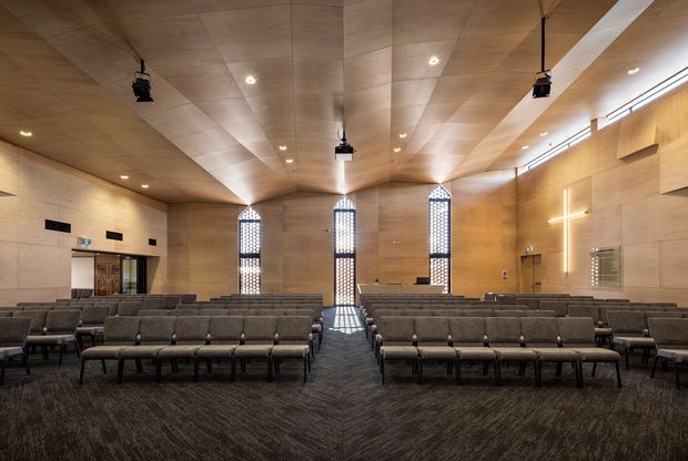 A large contemporary worship or assembly hall viewed from the seating area, with rows of upholstered chairs arranged around a central aisle. Warm timber wall and ceiling paneling, tall narrow patterned windows, recessed lighting, suspended projectors, dark carpet, and a simple illuminated cross create a refined, calm interior.