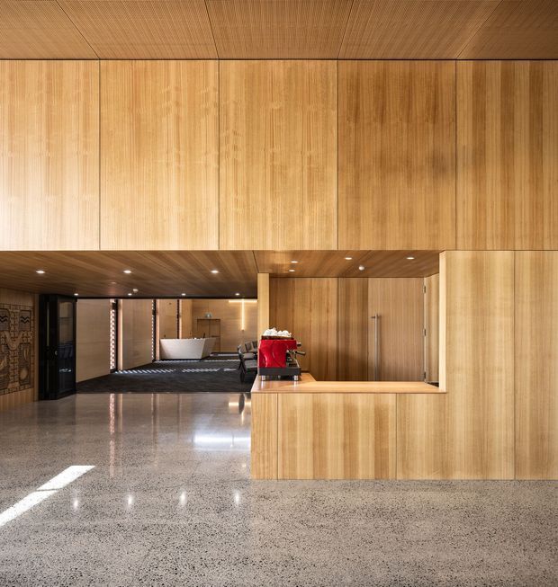 A refined contemporary commercial reception or hospitality lobby with extensive warm timber wall and ceiling panelling, integrated timber cabinetry, and a polished terrazzo floor. A reception counter with a red espresso machine occupies the foreground, while a lounge and service area extend into the background. Recessed ceiling spotlights and concealed lighting create a warm, polished atmosphere.