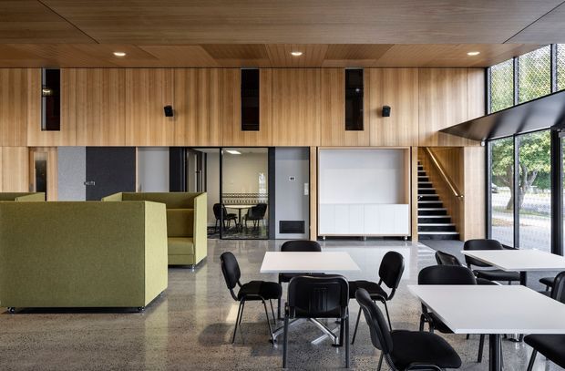 A contemporary communal interior with a dining and breakout area furnished with white rectangular tables and black chairs, alongside olive-green upholstered booth seating. The room features extensive warm timber wall and ceiling lining, polished concrete or terrazzo-like flooring, black-framed glazing, recessed lighting, a large white presentation screen, and a stairway leading to an upper level. Daylight enters through full-height windows overlooking greenery.