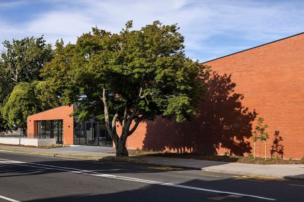 Exterior view of a low-rise contemporary brick building beside a street, with a long red-brick wall, a recessed glazed entrance, mature trees, paved footpath and road markings. The building has a simple angular roofline and restrained institutional or commercial character.