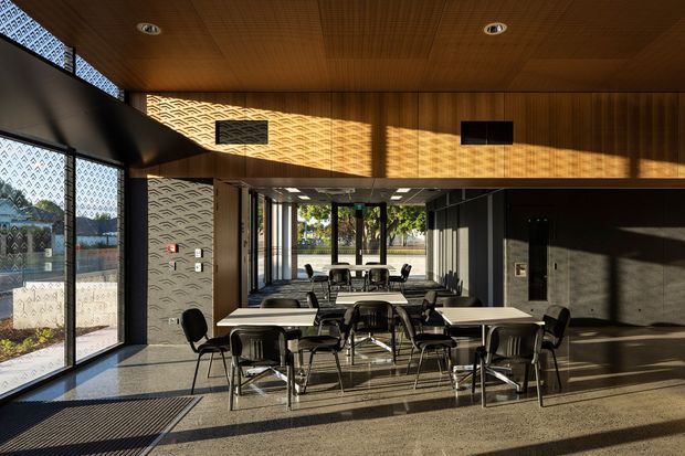 A contemporary commercial interior, likely a communal dining or meeting area, with multiple rectangular tables and black chairs arranged across a spacious room. Floor-to-ceiling glazing lines the left side, while patterned perforated screens, dark wall panels, and extensive warm timber ceiling panelling define the architecture. Strong late-afternoon sunlight creates patterned shadows across the walls and floor.