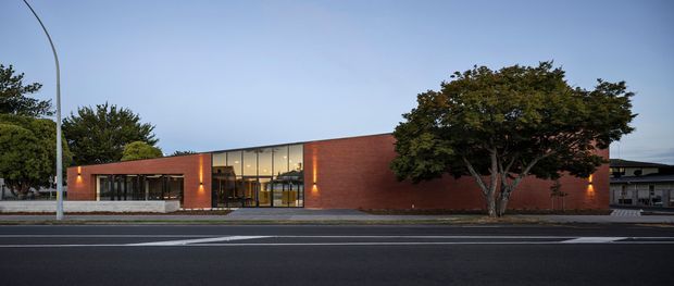Single-storey contemporary brick building viewed from across a street at dusk. The long, low facade combines warm red brick walls with a broad central band of floor-to-ceiling glazed doors and windows, framed in dark metal. Minimal exterior lighting illuminates the entrances and brickwork, with mature trees partly screening the right side. The image primarily presents the building exterior and streetscape.
