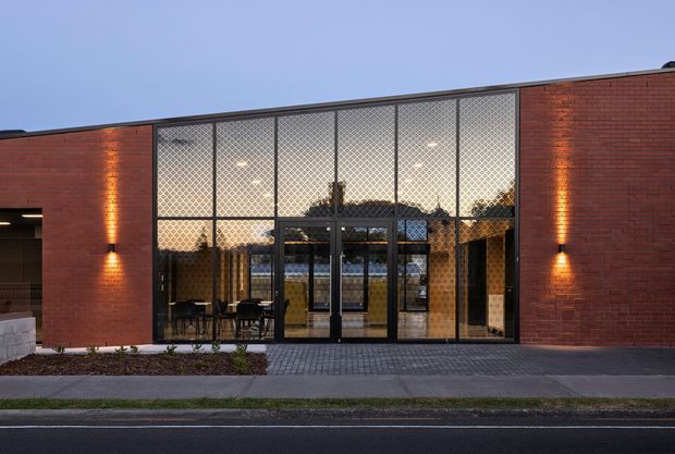 A low, contemporary brick building facade photographed at dusk. The symmetrical elevation features broad floor-to-ceiling glazed doors and windows with a distinctive patterned screen or mesh, warm interior illumination, and vertical wall sconces set into red brick walls. A paved pedestrian entry strip and road occupy the foreground.