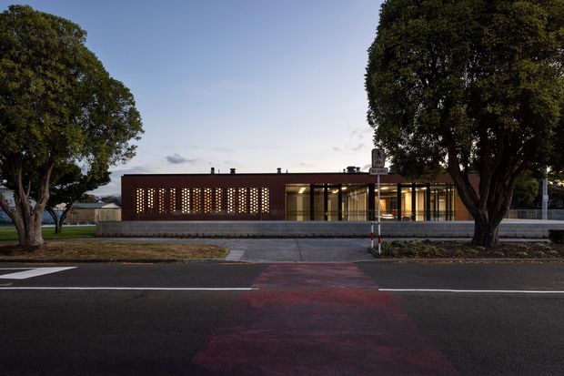 Street-facing view of a low-rise contemporary building at dusk, framed by mature trees. The facade combines a dark brick screen with regularly spaced perforations and a glazed entrance or frontage beneath a flat roof. A paved roadway and pedestrian crossing occupy the foreground.