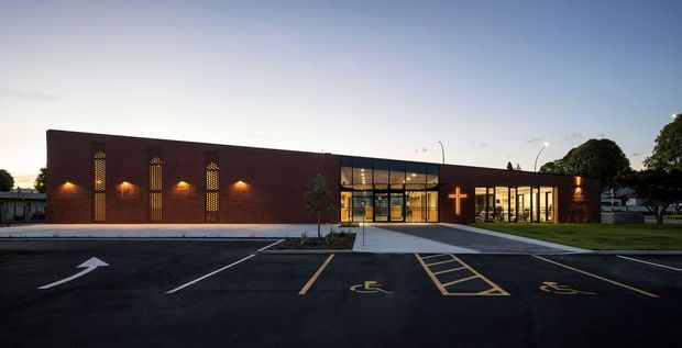 Exterior architectural photograph of a low, contemporary brick community or commercial building at dusk. The long dark brick facade includes narrow decorative perforated screens and wall lighting, with a glazed central entrance and a bright cross-shaped feature visible on the right-hand facade. A paved parking area and accessible parking markings occupy the foreground, with lawns and mature trees around the building.