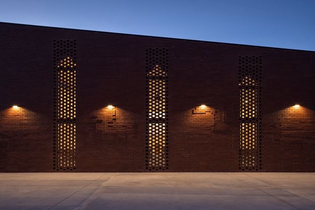 A dark brick building facade photographed at dusk, featuring three tall, narrow arched openings with perforated brick screens and warm light glowing from within. Small wall-mounted lights illuminate the textured brickwork, while a broad paved forecourt occupies the foreground beneath a clear blue sky.