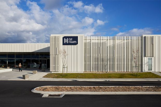 Exterior view of a contemporary single-storey healthcare facility with a long, rectilinear form. The facade combines extensive vertical white fins or battens with dark floor-to-ceiling glazing at the entrance, and a prominent QE Health sign. A paved forecourt, planted strip, and newly finished roadway are visible in front under a partly cloudy sky.