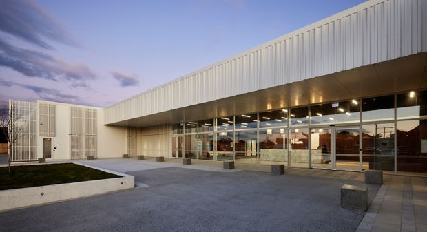 Exterior view of a contemporary low-rise commercial building at dusk, featuring a long cantilevered white vertically ribbed metal facade, recessed soffit lighting, extensive floor-to-ceiling glazed frontage, and a paved forecourt with simple concrete seating blocks. A smaller matching service volume and landscaped planter are visible to the left.
