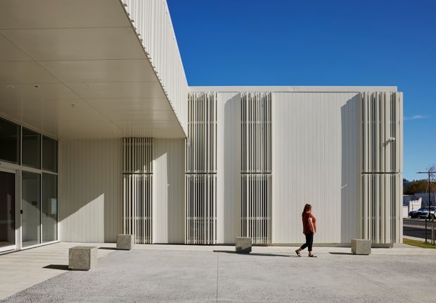 Contemporary white institutional or commercial building exterior with a broad projecting canopy, extensive glazed openings, and prominent vertical white fins screening the facade. The minimalist courtyard or forecourt is paved in pale concrete with simple square concrete seats or bollards, while a person provides scale. Bright clear daylight emphasizes the crisp geometric forms.