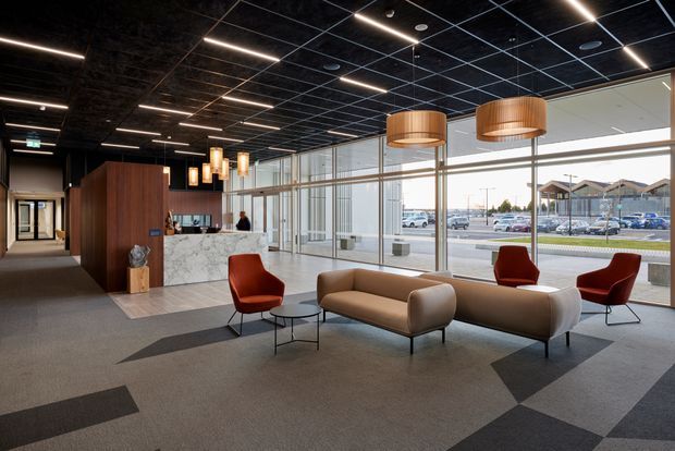 A contemporary commercial reception and waiting area with expansive floor-to-ceiling glazing, a marble reception desk, timber feature joinery, sculptural upholstered sofas and red lounge chairs. The space has a dark exposed grid ceiling with linear lighting and large woven pendant lights, while patterned carpet tiles define the open seating zone. A car park and neighbouring buildings are visible outside.