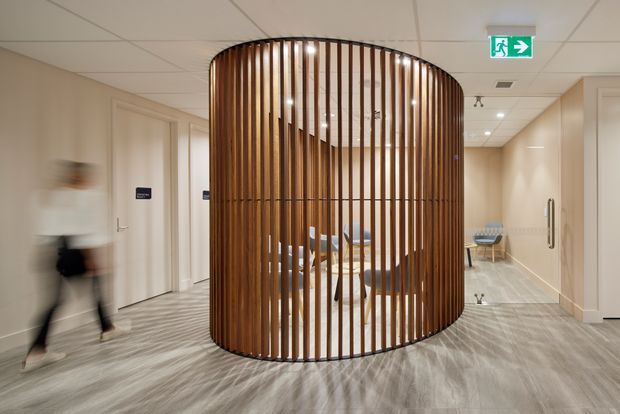 Interior view of a contemporary commercial hallway or lobby featuring a prominent curved cylindrical timber slat partition enclosing a seating area. The corridor has pale beige walls, grey patterned carpet, a suspended acoustic tile ceiling with recessed lighting, and accessible circulation around the sculptural screen. An emergency exit sign and institutional doors reinforce the commercial setting.