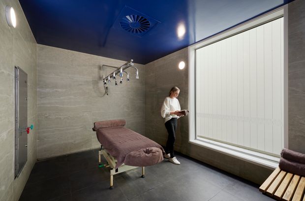 A contemporary treatment or massage room with a padded treatment table, wall-mounted shower fixtures, large obscured window, dark tiled floor, stone-look wall surfaces, and a glossy deep-blue ceiling. A timber slatted bench with cushions sits beside the window. The space is brightly lit and clinically refined, resembling a spa, wellness facility, or commercial therapy room.