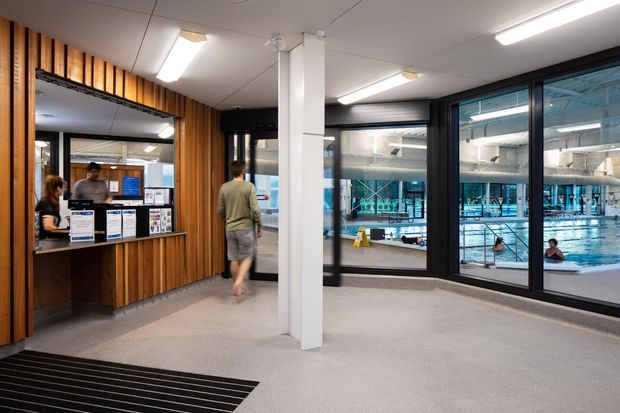 Interior view of a modern aquatic centre reception and circulation area. A timber-lined reception counter sits to the left, while large black-framed glazed partitions provide views into an indoor swimming pool. The space has a light grey carpeted floor, white ceiling panels, exposed white structural columns, and linear fluorescent lighting. People and pool activity are visible beyond the glazing.
