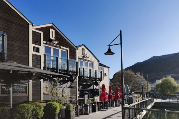 Exterior streetscape of a multi-storey waterfront-style commercial building with dark timber cladding, pale trim, projecting glazed balconies, and ground-floor restaurant or hospitality spaces. A pedestrian promenade with outdoor umbrellas, railings, and a distinctive street lamp runs alongside the building, with a wooded mountain backdrop visible beyond.