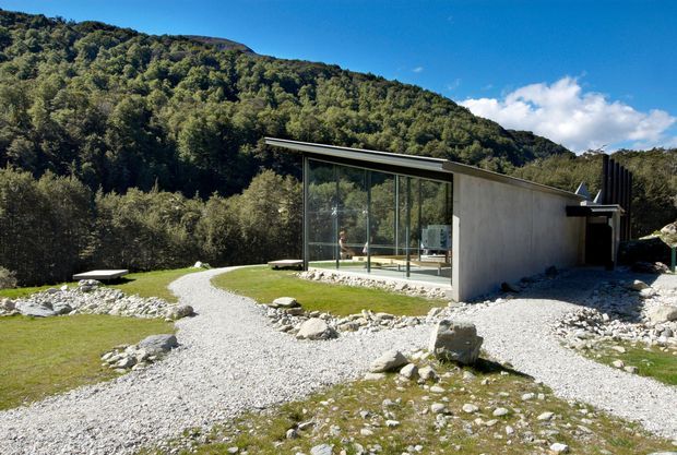 A minimalist single-storey modern building set in a mountainous, heavily wooded landscape. The structure has a long low sloping roof, a solid concrete or rendered side wall, extensive floor-to-ceiling glazing, and a gravel path leading through a sparse rocky lawn. The surrounding terrain and vegetation dominate the setting.