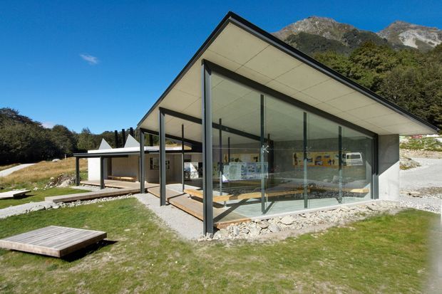 A low-slung contemporary pavilion-like building set against a dramatic alpine mountain backdrop. The structure has a sharply pitched asymmetrical roof, extensive floor-to-ceiling glazing with dark slender frames, pale soffits, and open-plan interior spaces visible through the glass. Gravel, lawn, timber platforms, and simple outdoor elements surround the building.