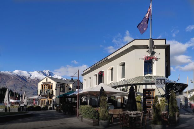 A sunny streetscape showing a two-storey commercial pub building with a light rendered facade, arched windows, dark trim, and a glazed street-front dining terrace. Outdoor tables, umbrellas, signage, and flags line the pedestrian area, with snow-capped mountains visible in the background.