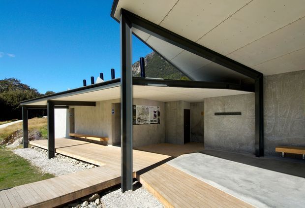 Low-slung contemporary building with broad cantilevered roof planes, exposed black structural posts and beams, and extensive triangular and rectangular glazing. The image shows an open covered entry or terrace with concrete walls and floor areas, timber decking and walkways, built-in timber benches, and a gravel landscape. A mountain slope is visible beyond the glazing and roofline.