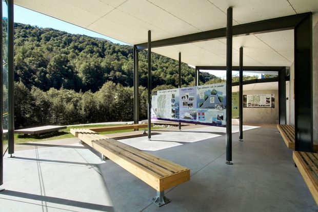An open-air covered pavilion or public exhibition space set against a densely wooded hillside. The structure has a flat, light-coloured ceiling, slender dark steel posts and beams, polished concrete flooring, and extensive timber bench seating arranged around the perimeter. Educational display boards occupy the centre and rear walls, while large openings frame the surrounding landscape. The architecture is restrained and contemporary, with strong indoor-outdoor connection and abundant natural light.