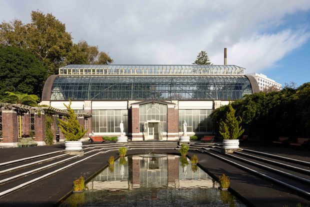 Exterior view of a grand historic glasshouse or conservatory, featuring a large glazed curved roof and facade framed by brick piers and pale stone detailing. Formal symmetrical steps, clipped hedges, urn planters, classical statues, benches, and a long reflecting pool create a landscaped ceremonial forecourt.