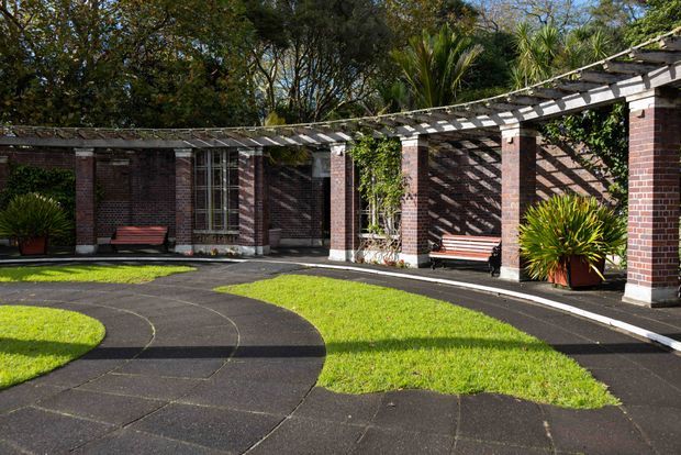 A landscaped courtyard beside a low brick building, featuring a long open timber pergola supported by brick columns. Curved dark paving paths divide bright green lawn areas, with benches, potted plants, and tall windows along the building facade. Strong sunlight creates patterned shadows across the paving and brickwork.