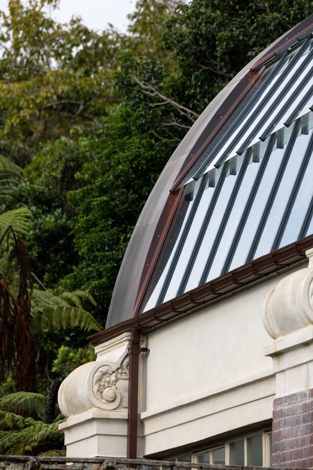 Close-up exterior architectural detail of a heritage-style building set among dense greenery. The image shows an ornate cream rendered parapet with carved scroll ornamentation, dark reddish-brown metal guttering and trim, brickwork, and a steeply curved glazed roof or skylight structure with dark framing.