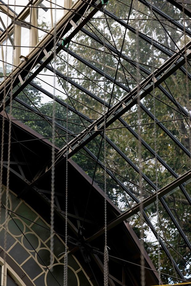 A dramatic upward-looking view inside a glazed, greenhouse-like architectural structure. The frame is dominated by a complex black-painted metal roof and truss system with extensive glass panels, suspended chains, and diagonal bracing. A dark pitched roof volume and decorative patterned wall panel are visible below, while mature trees show through the glazing. The image emphasizes exposed structural steel, transparent enclosure, and a tall, airy overhead space.