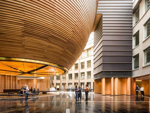 A grand contemporary commercial interior atrium or lobby with a dramatic curved timber-slat ceiling and integrated amber-lit circular canopy above a reception or seating area. Tall glazed and metal-clad building volumes surround a polished dark floor, with cylindrical timber-clad columns, abundant daylight, and people using the spacious public circulation zone.