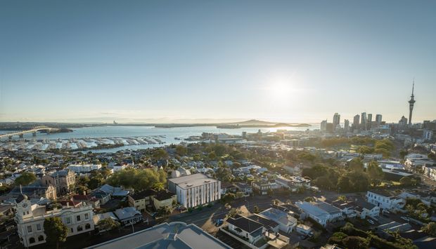 Aerial panoramic view over a dense urban waterfront, showing a marina and harbour in the middle distance, low-rise residential neighbourhoods in the foreground, and a prominent city skyline with a tall observation tower on the right. The scene is photographed in warm low-angle sunlight under a clear sky.
