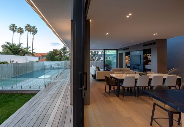 A contemporary open-plan living and dining area viewed through fully opened floor-to-ceiling glazing, connected to a timber deck and glass-sided swimming pool. The interior features warm timber flooring, dark built-in cabinetry, a large dining table, upholstered dining chairs, a lounge seating area and recessed ceiling lighting. Palm trees and neighbouring residential buildings are visible beyond the pool.