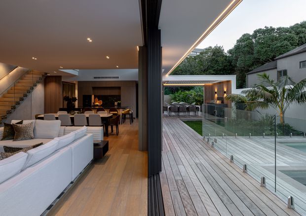 A sophisticated open-plan living and dining area opening directly onto a large timber deck and pool terrace. The interior features pale upholstered sofas, a dining table, dark timber wall panelling and cabinetry, recessed lighting, and a timber staircase. Full-height retractable glazed doors create a seamless connection to the outdoor entertaining area, which includes glass pool fencing, an outdoor dining/lounge zone, and lush planting. The design has a refined contemporary character with warm timber finishes and strong indoor-outdoor flow.