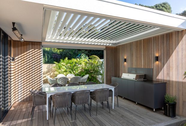 A covered outdoor dining and entertaining area with a timber deck, timber-lined walls, slatted outdoor dining furniture, and a built-in barbecue cabinet. A retractable louvred roof provides filtered sunlight, while the open end looks onto landscaped greenery.