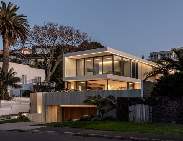 Twilight exterior view of a contemporary multi-level residence in an established suburban setting. The house features strong horizontal lines, extensive floor-to-ceiling glazing, a projecting upper storey, warm integrated lighting, dark vertical cladding, a timber garage door, and a low dark-stone boundary wall. Palm trees and neighbouring homes frame the property.