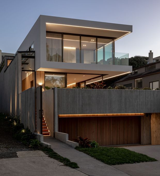 Twilight exterior view of a contemporary multi-level urban residence with a crisp cantilevered upper volume, extensive floor-to-ceiling glazing, recessed linear lighting, dark vertical cladding, and a timber-lined garage integrated into the lower level. A high solid boundary wall, landscaped entry stair, balcony glazing, and neighbouring homes frame the facade.