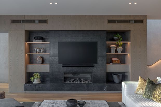 Contemporary living room media wall with a wall-mounted television, linear fireplace, dark stone feature surround, and light timber built-in cabinetry with illuminated open shelving. A white sofa and marble-look coffee table sit in the foreground, creating a refined, warm-toned lounge.