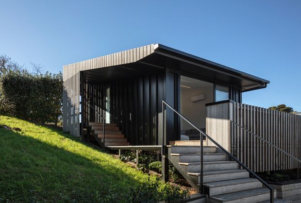 Exterior view of a compact contemporary elevated dwelling on a sloping grassy site. The building has dark vertical cladding, a shallow flat roof with broad overhangs, large glazed openings, and a timber-clad balustrade enclosing an outdoor access deck. External stairs with a simple metal handrail lead to the raised entry.