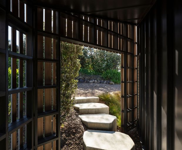 A dark, narrow architectural passage or entry enclosure opens onto a landscaped garden path. The enclosure features black metal framing with vertical timber slats, framed glazing or open panels, and a dark ceiling. Large irregular pale concrete stepping stones lead through mulched planting toward a low stone retaining wall and dense greenery. Strong contrast between the shaded interior and sunlit garden creates a dramatic, refined transition space.