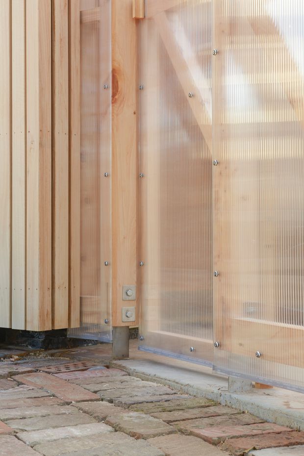 Close-up exterior architectural detail showing vertical natural timber cladding beside translucent corrugated polycarbonate panels fixed with exposed metal fasteners. A timber-framed translucent door or screen reveals structural framing behind it. The base meets an uneven reclaimed brick-paved surface and concrete threshold.