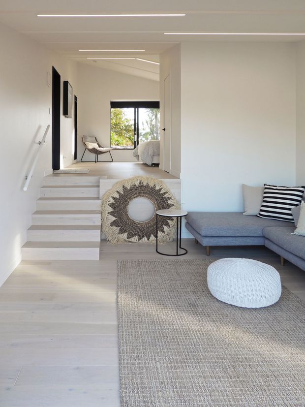 Bright, minimalist open-plan living area with pale timber flooring, a grey sectional sofa, woven neutral rug, round knitted ottoman, and black-and-white striped cushion. A short flight of timber stairs leads to a raised bedroom area, with a lounge chair and bed visible beyond. Slim linear recessed ceiling lights and white walls create a clean, airy interior.