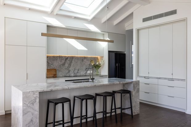 Bright contemporary kitchen with a large marble-look island, four black bar stools, white full-height cabinetry, integrated appliances, a matching stone splashback, and a linear timber pendant. Multiple skylights and white structural beams create an airy, high-ceilinged space, while dark flooring provides contrast.