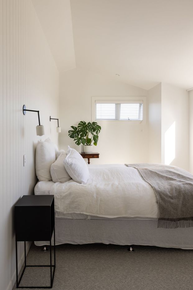Bright, compact bedroom with a neatly made bed against a white vertical-panel feature wall. The room includes minimalist black wall-mounted reading lamps, a slim black bedside table, white bedding, a grey throw, soft grey carpet, a small wooden console with a potted monstera, and a horizontal window with white blinds. The overall look is simple, airy, and contemporary with natural accents.