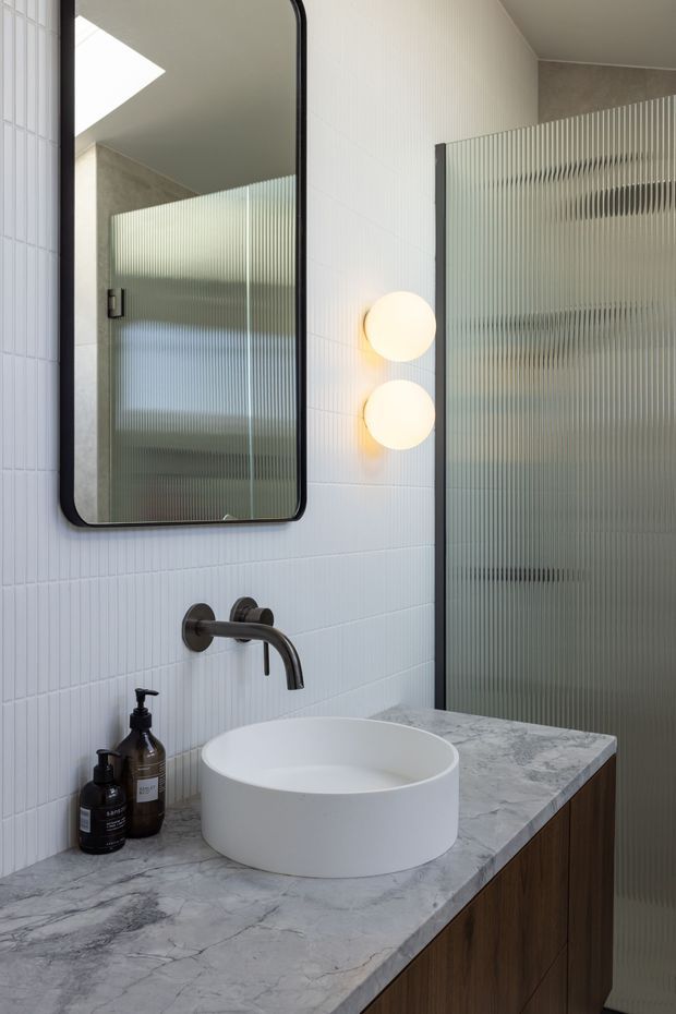 A refined contemporary bathroom vanity featuring a round white vessel basin on a grey-veined stone benchtop, dark timber cabinetry, wall-mounted dark metal tapware, vertically ribbed white wall tiles, and a large rounded-corner mirror. A fluted translucent glass shower screen and two spherical wall lights add texture and softness.