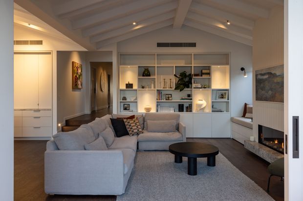 A refined contemporary living room with a large grey sectional sofa, round dark coffee table, built-in illuminated shelving and cabinetry, and a linear fireplace set into a paneled feature wall. The room has a warm neutral palette, dark timber flooring, a vaulted ceiling with exposed painted beams, recessed lighting, and an open hallway leading toward the entry or adjoining spaces.