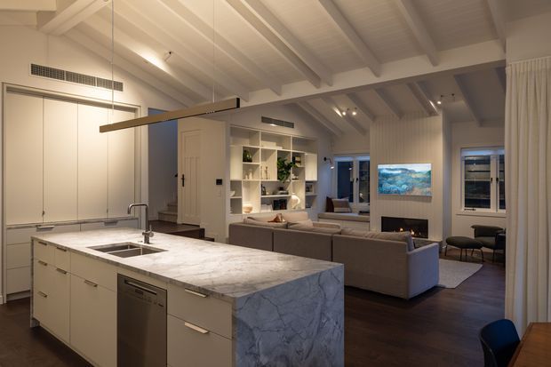 Open-plan contemporary interior showing a kitchen island with a large pale veined stone benchtop, integrated cabinetry and sink, opening into a living room. The living area has dark timber flooring, a sectional sofa, built-in shelving, a fireplace, and exposed painted ceiling beams with sloped vaulted forms. Warm recessed and pendant lighting creates a refined, inviting atmosphere.